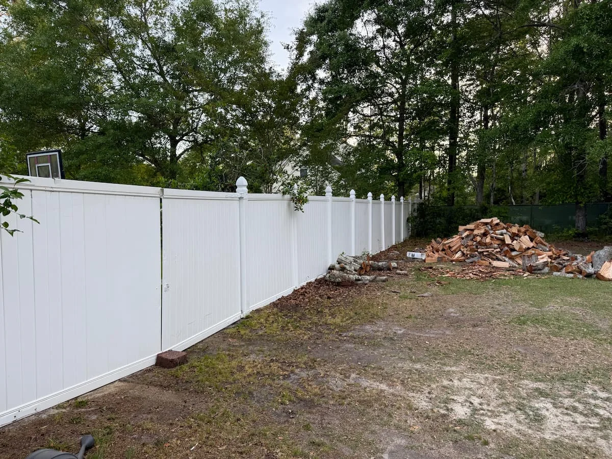 A pressure-washed white vinyl fence showcasing a clean, bright finish against a backdrop of natural greenery and woodpile debris.